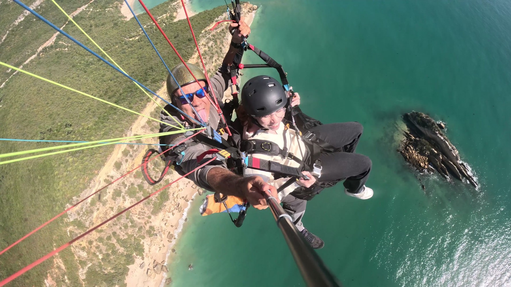 Tandem paraglider over turquoise water with visible coastline and rocks.