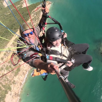 Tandem paraglider over turquoise water with visible coastline and rocks.