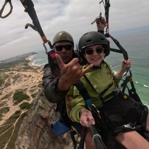 Two people paragliding over a coastal landscape with ocean and cliffs visible.