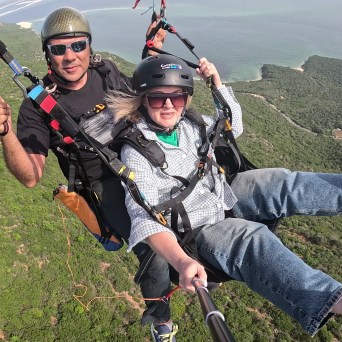 Two people paragliding over green hills with a view of the ocean.