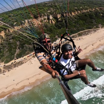 Two people paragliding over a scenic beach and cliffs with green foliage.