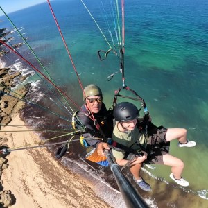 Two people paragliding over a coastline with clear blue sea and rocky cliffs.