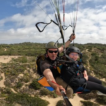 Two people paragliding over a scenic landscape with trees and sand dunes visible below.