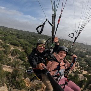 Two people paragliding over a green landscape with trees, capturing a selfie mid-air.