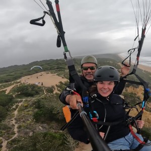 Two people tandem paragliding over a coastal landscape with cloudy skies.