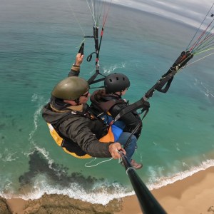 Tandem paragliders flying over a beach with clear blue water.