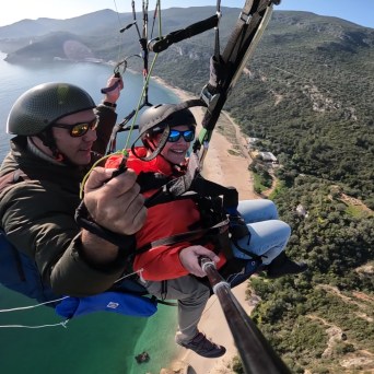 Two people paragliding over a coastal landscape with mountains and sea.