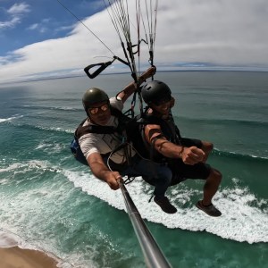 Two people tandem paragliding over ocean waves, one holding a selfie stick.