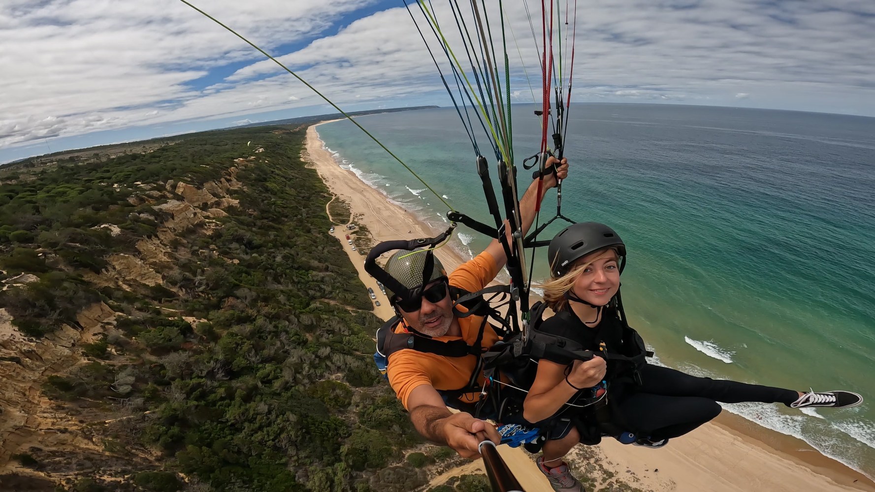 Two people paragliding over a coastal landscape with beach and ocean view.
