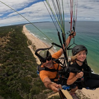 Two people paragliding over a coastal landscape with beach and ocean view.