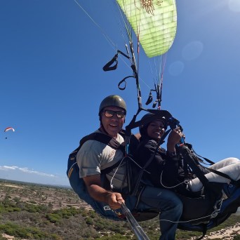 Two people paragliding over a landscape with clear blue sky and distant paraglider.