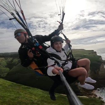 Two people tandem paragliding over a coastal cliff.