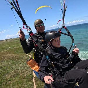 Two people tandem paragliding over a coastal landscape with blue ocean and clear sky.