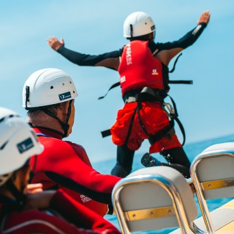 Person in red gear jumping off a boat, others watching, all wearing helmets.