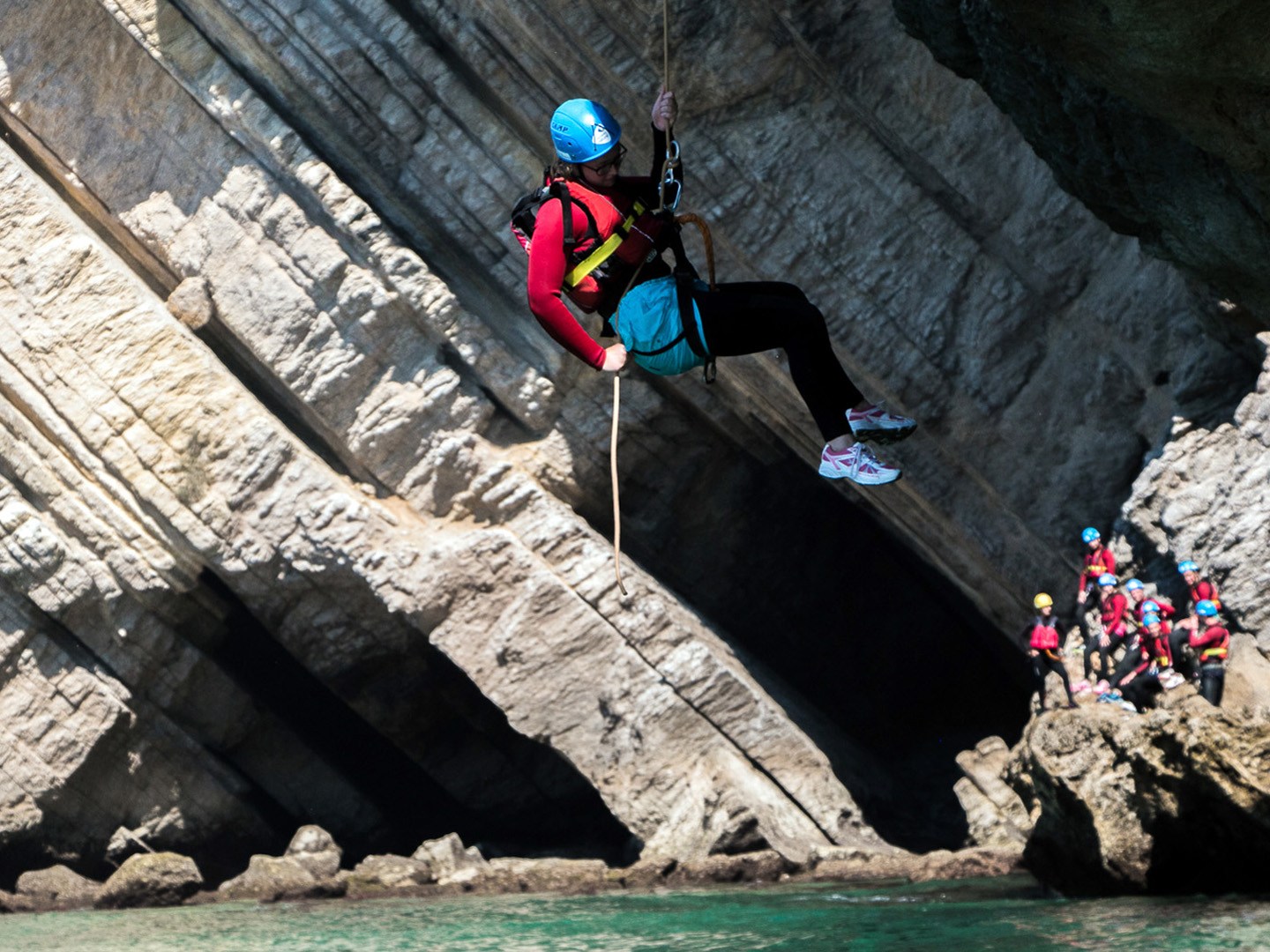 Person in helmet and harness swings from rope in rocky coastal area, with group watching nearby.