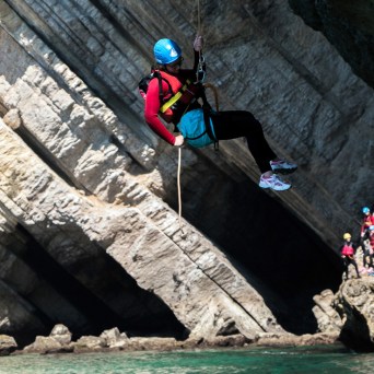 Person in helmet and harness swings from rope in rocky coastal area, with group watching nearby.