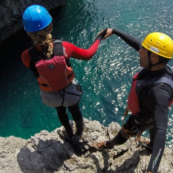 Two people in helmets and life vests stand on a cliff edge above water, holding hands.