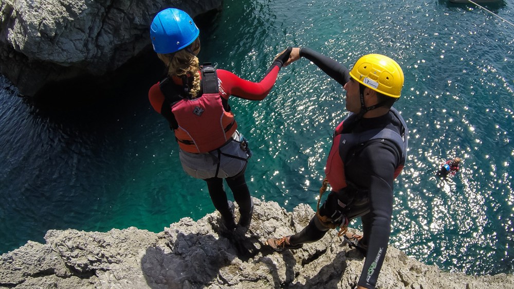 Two people in helmets and life vests stand on a cliff edge above water, holding hands.