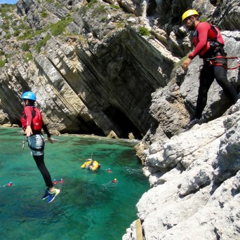 Person cliff jumping into turquoise water with others in life jackets below, rocky cliffs in background.