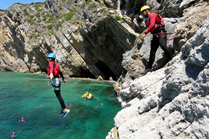 Person cliff jumping into turquoise water with others in life jackets below, rocky cliffs in background.