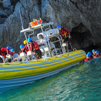 People in helmets and life jackets on a yellow boat, some swimming near rocky cliffs.