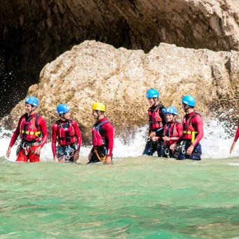 Group of people in helmets and wetsuits walking in water near a rocky cave entrance.