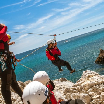 Group of people in helmets and harnesses zip-lining over rocky coast with ocean in background.