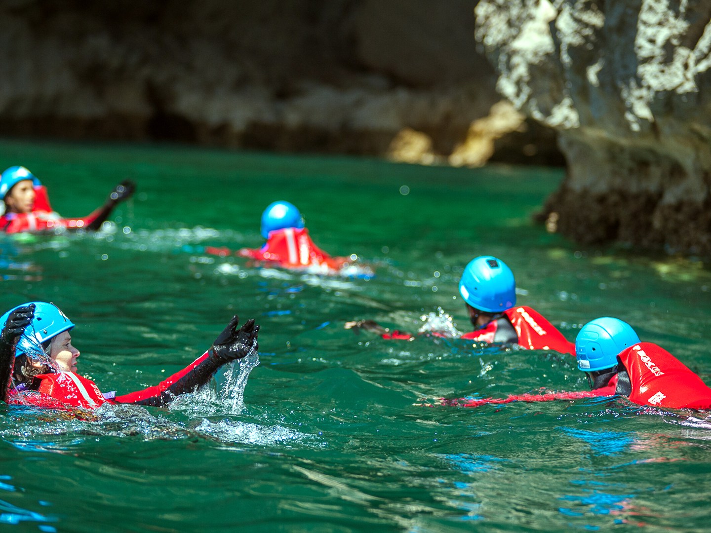 Group of people in red suits and blue helmets swimming in a clear green water cave.