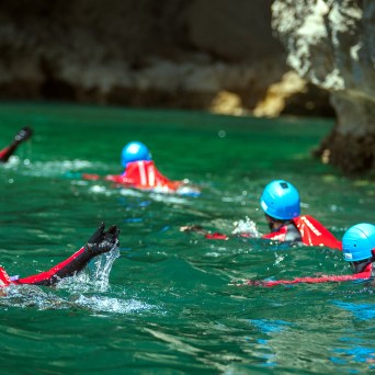 Group of people in red suits and blue helmets swimming in a clear green water cave.