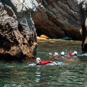 People swimming near large rocks wearing helmets and wetsuits in clear water.