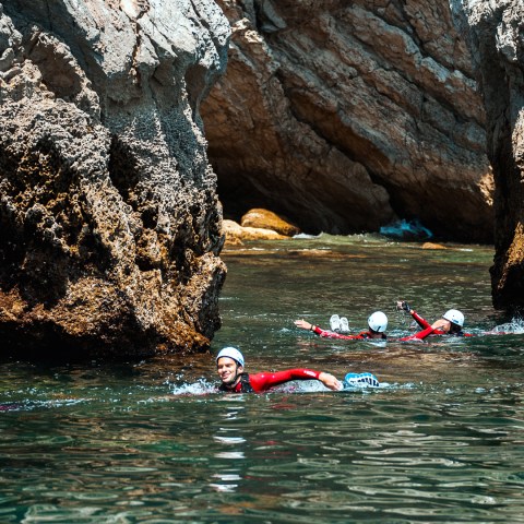 People swimming near large rocks wearing helmets and wetsuits in clear water.