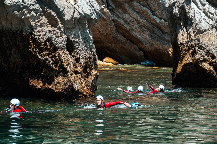People swimming near large rocks wearing helmets and wetsuits in clear water.