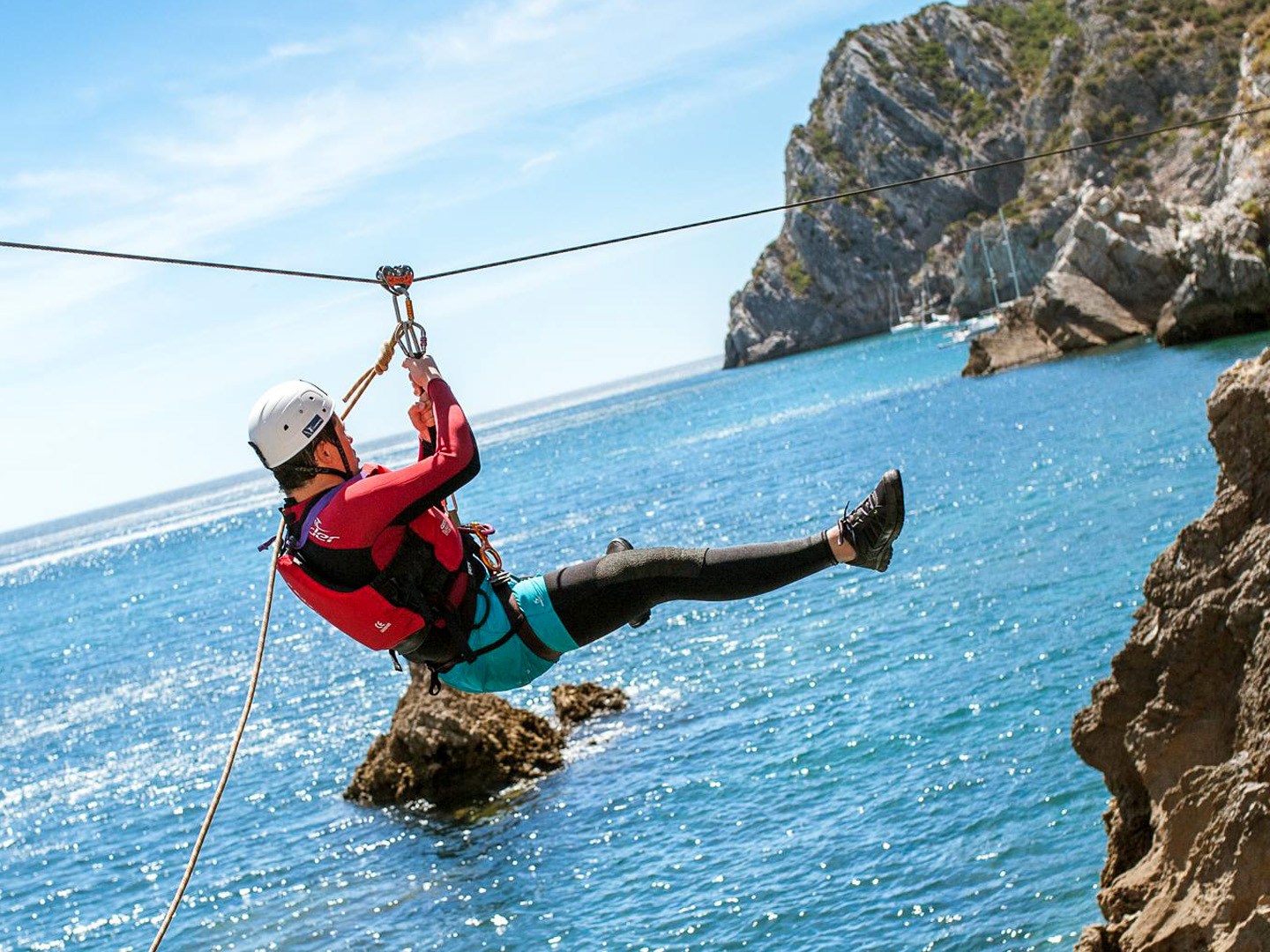 Person ziplining over water near rocky coastline on a sunny day.
