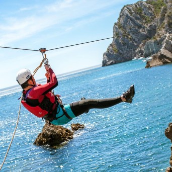 Person ziplining over water near rocky coastline on a sunny day.