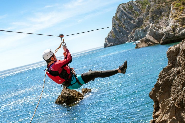 Person ziplining over water near rocky coastline on a sunny day.