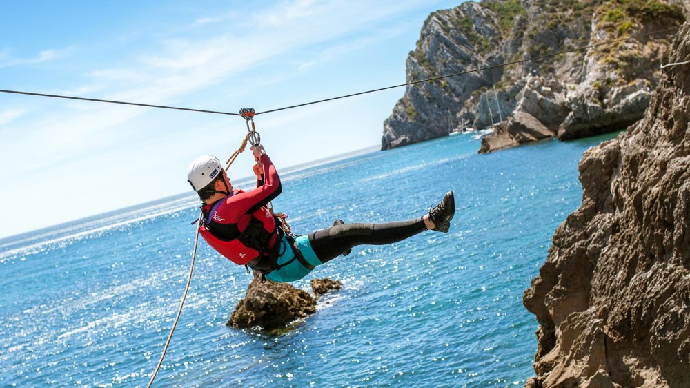 Person ziplining over water near rocky coastline on a sunny day.