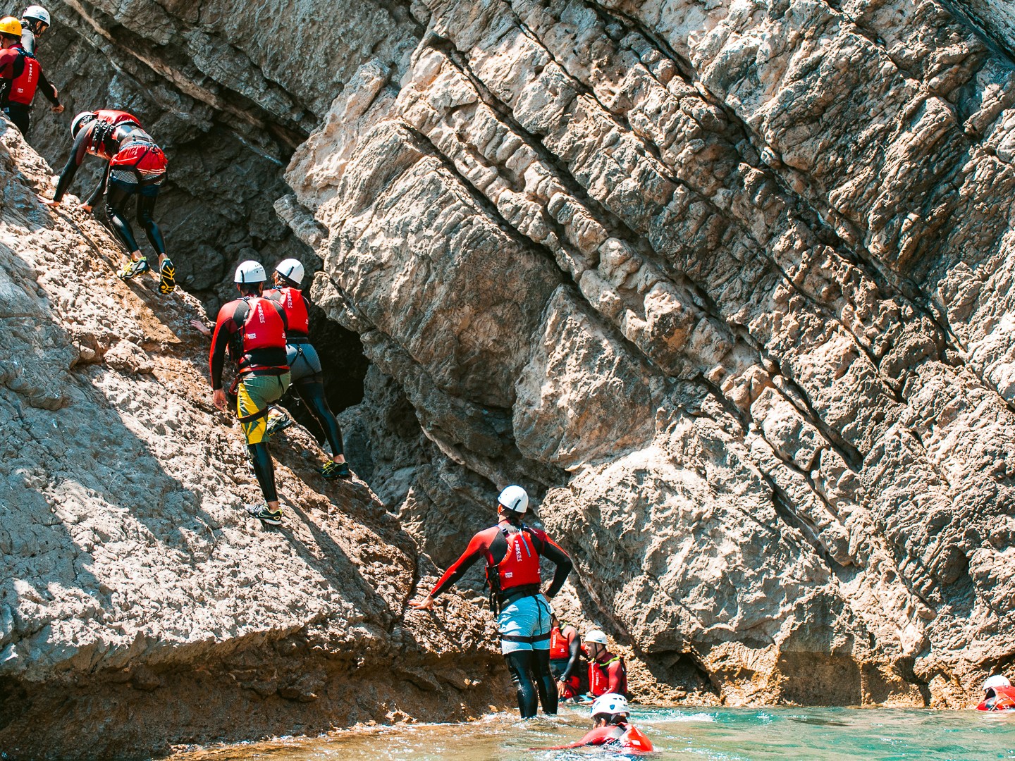 Group in red gear and helmets climbing rocky cliff by water.