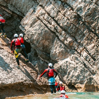 Group in red gear and helmets climbing rocky cliff by water.
