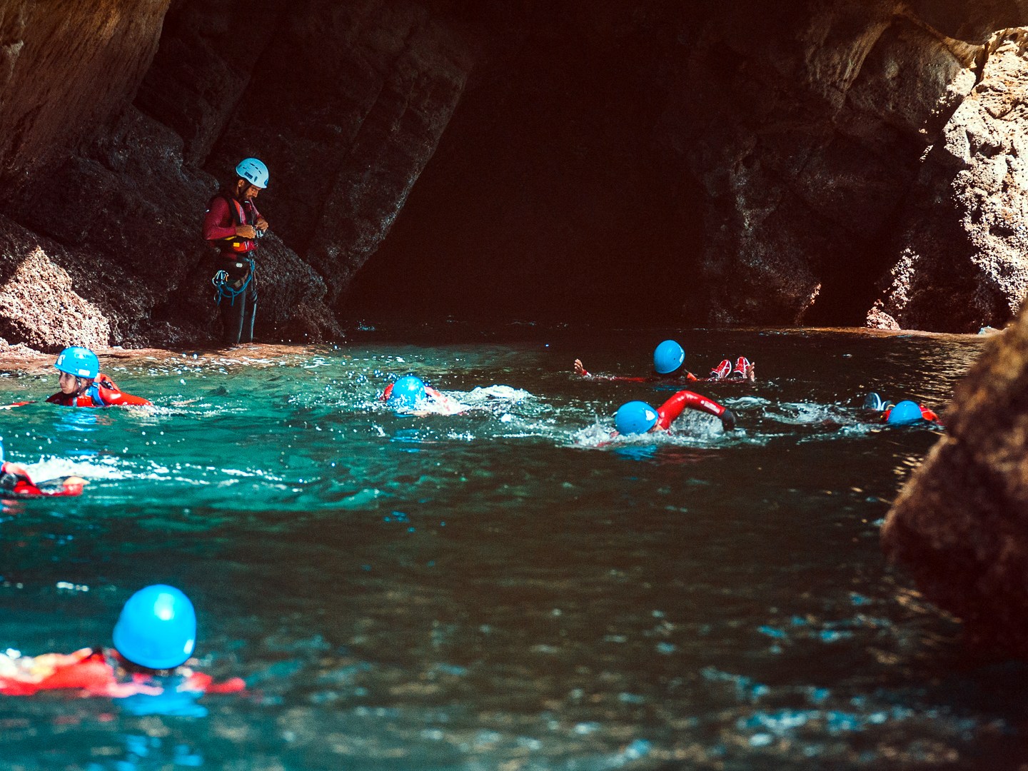 People wearing helmets and life jackets swim near a rocky cave entrance.