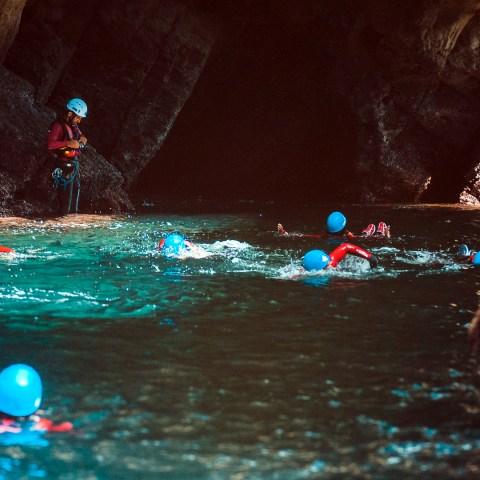 People wearing helmets and life jackets swim near a rocky cave entrance.