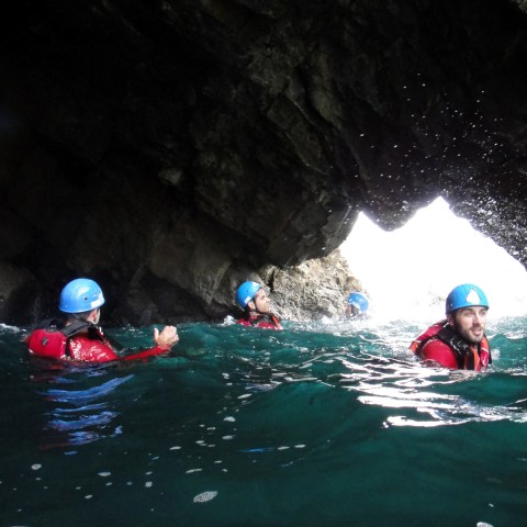 People with helmets swimming in a sea cave with rocky walls.