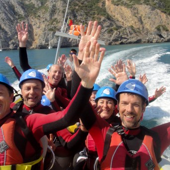 Group of people in helmets on a boat, smiling and raising hands, rocky coastline in background.