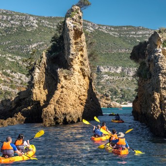 Kayakers in orange boats explore rocky coastal formations under a clear blue sky.