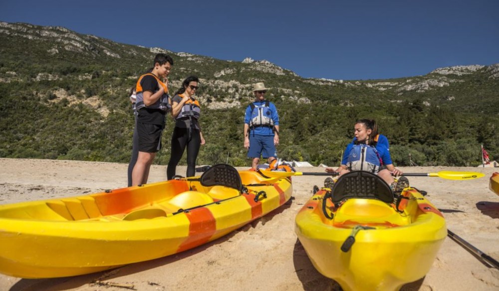Four people in life vests preparing kayaks on a beach with mountains in the background.