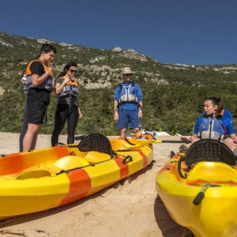 Four people in life vests preparing kayaks on a beach with mountains in the background.