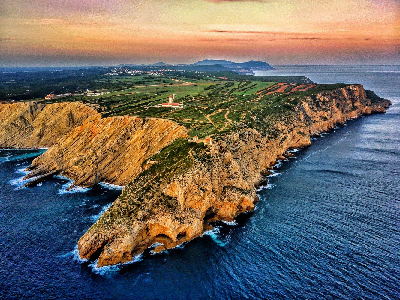 Aerial view of a rocky coastline with cliffs and a lighthouse at sunset.