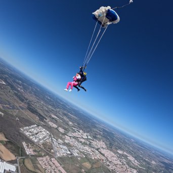 Tandem skydivers with open parachute above a landscape, under a clear blue sky.