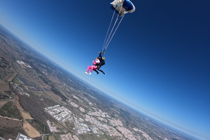 Tandem skydivers with open parachute above a landscape, under a clear blue sky.