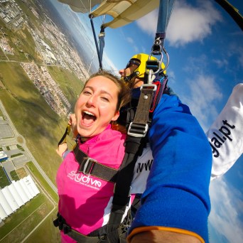 Two people tandem skydiving with a parachute open over a landscape, one smiling excitedly at the camera.