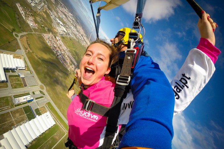 Two people tandem skydiving with a parachute open over a landscape, one smiling excitedly at the camera.